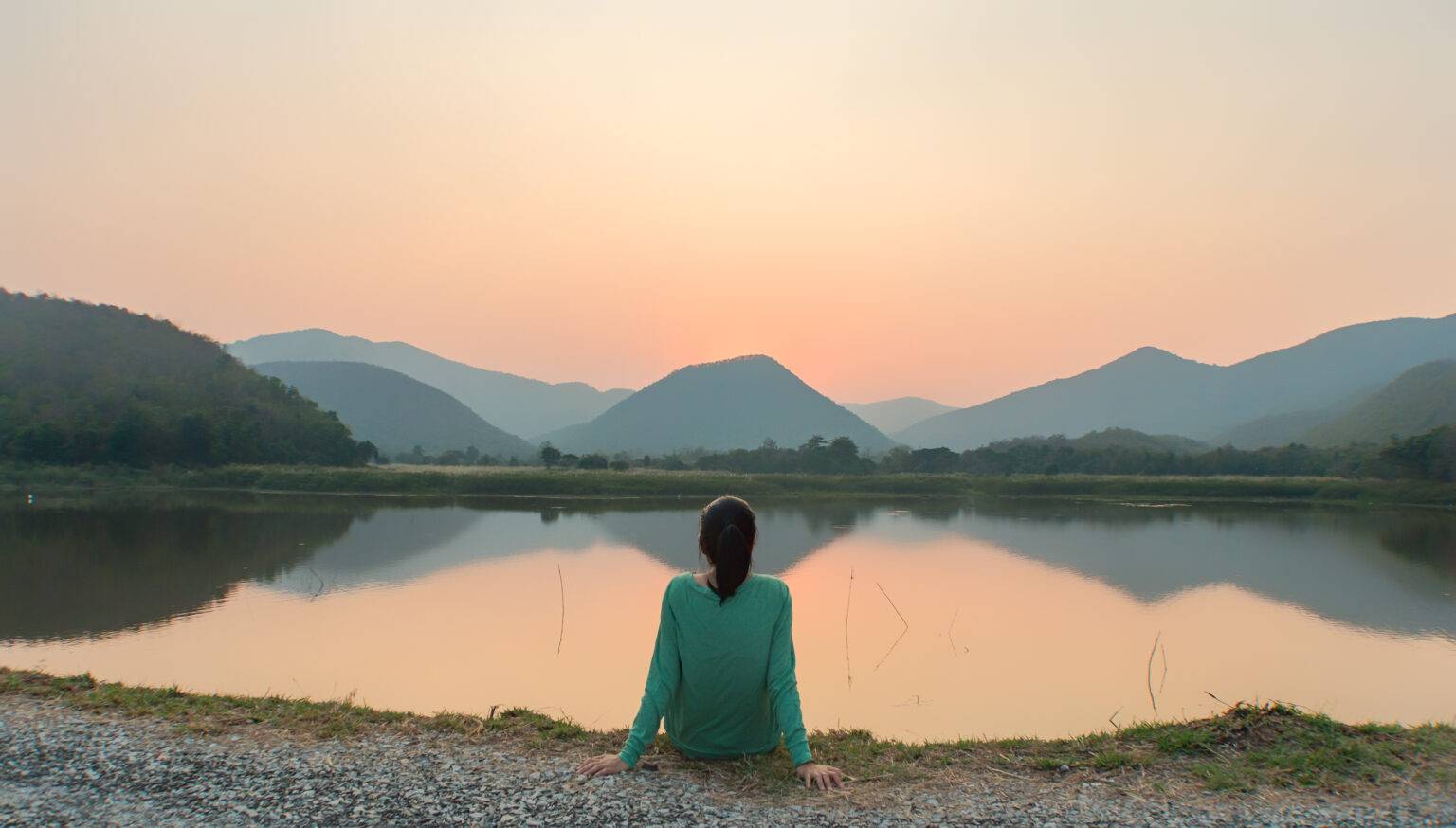 Woman sitting by a calm lake at sunset, looking out at the mountains