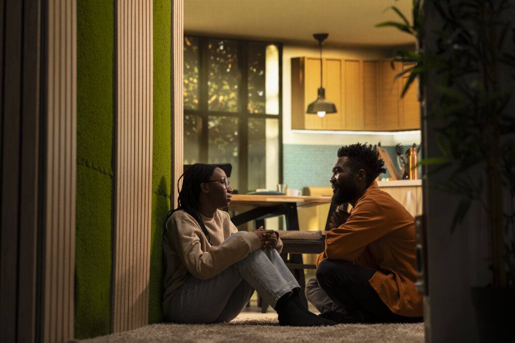Two friends sitting on the floor, facing each other and talking in a cozy room with a warm kitchen in the background.