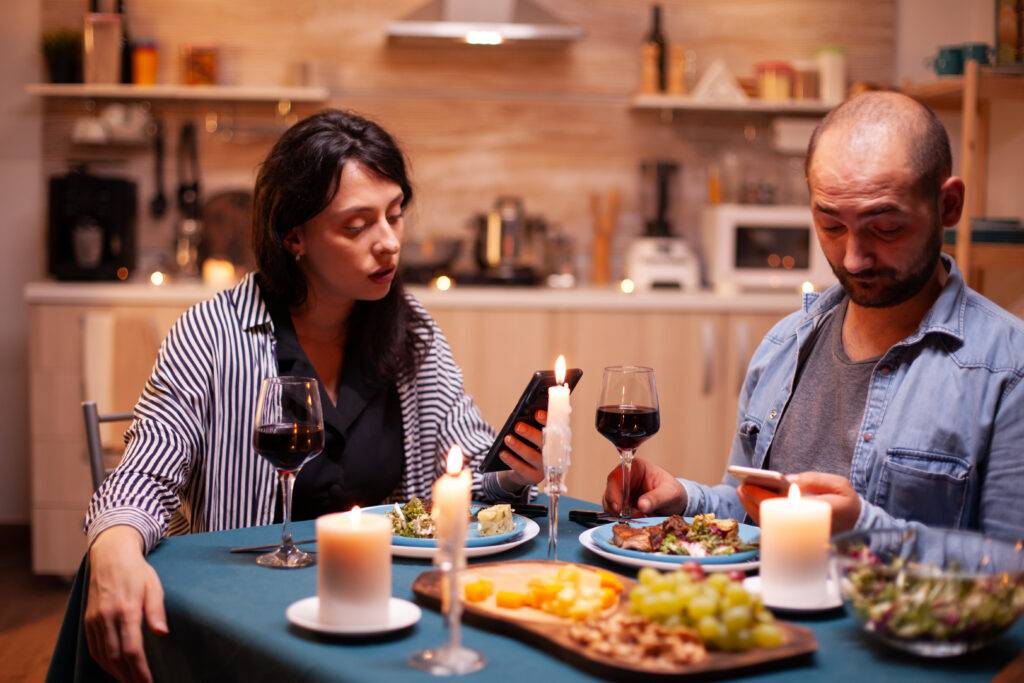 Two people dining at a candlelit table, looking at their phones amid plates and wine glasses in a cozy kitchen.