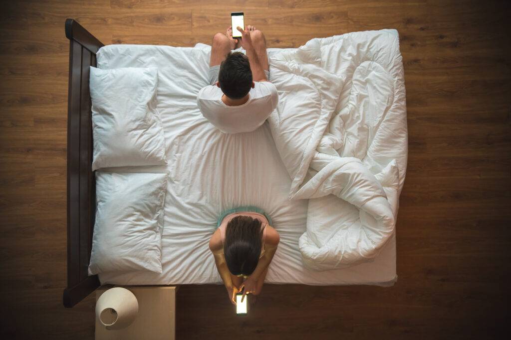 Overhead view of a couple lying in bed, each using a smartphone on white sheets.