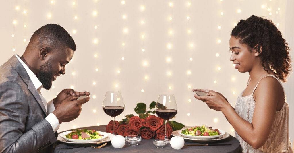 Black couple taking photos of meal in restaurant • Experience Hope Counseling Couple in formal attire sharing a romantic dinner at a table with wine glasses and a red rose centerpiece, fairy lights in the background.