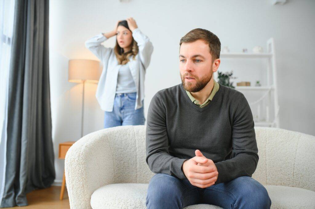 Bearded man in a dark sweater sits on a cream sofa, looking down with hands together, while a distressed woman stands behind him holding her head in a living room.