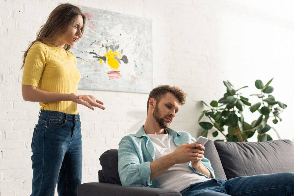 Woman in a yellow ribbed shirt stands and gestures as a man on a sofa looks at his phone in a bright, modern room with a plant nearby.
