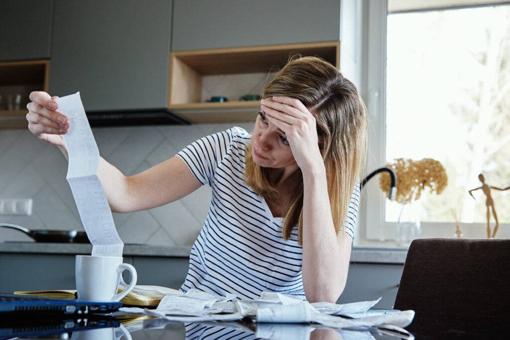 Woman sits at a kitchen table clutching a long receipt, looking stressed amid scattered papers and a mug on the table.