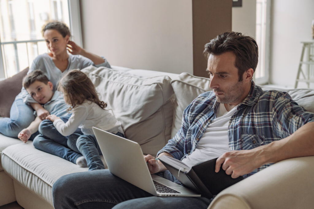 Man with a laptop sits on a couch to the right as a woman and two children cuddle on the left in a bright living room.