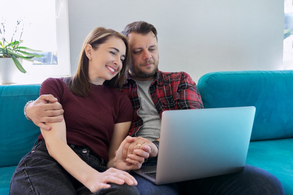 Smiling couple sitting close together on a teal sofa, holding hands and looking at a laptop screen.