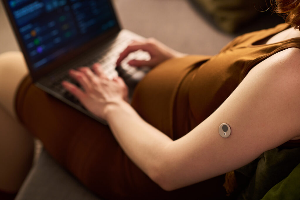 Person typing on a laptop while wearing a circular medical sensor on their forearm.