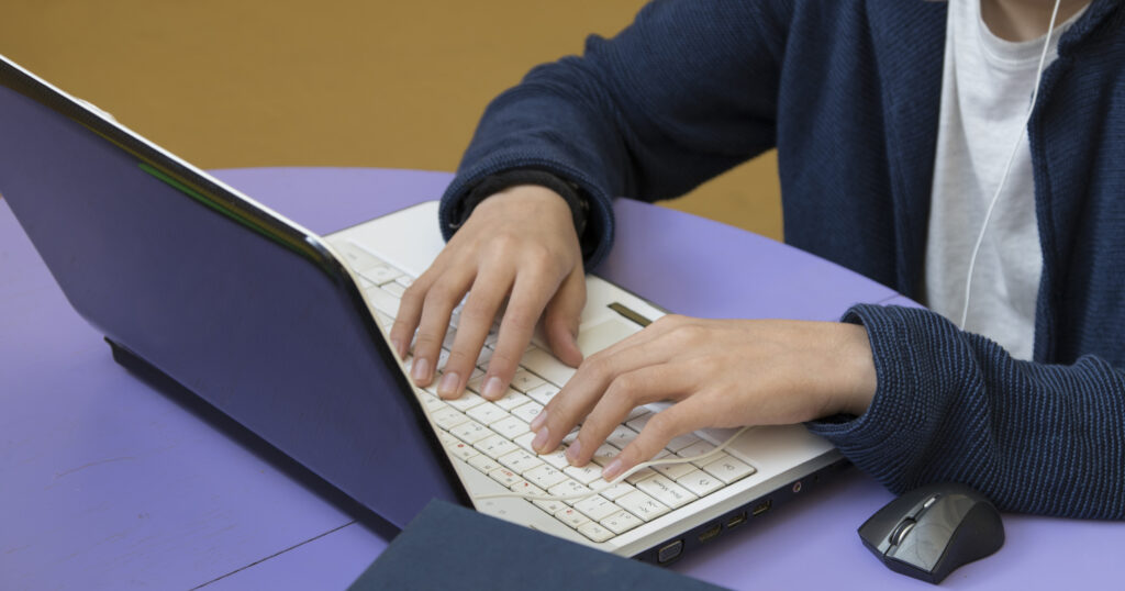 Person typing on a white laptop at a purple desk, wearing a blue cardigan/hoodie.